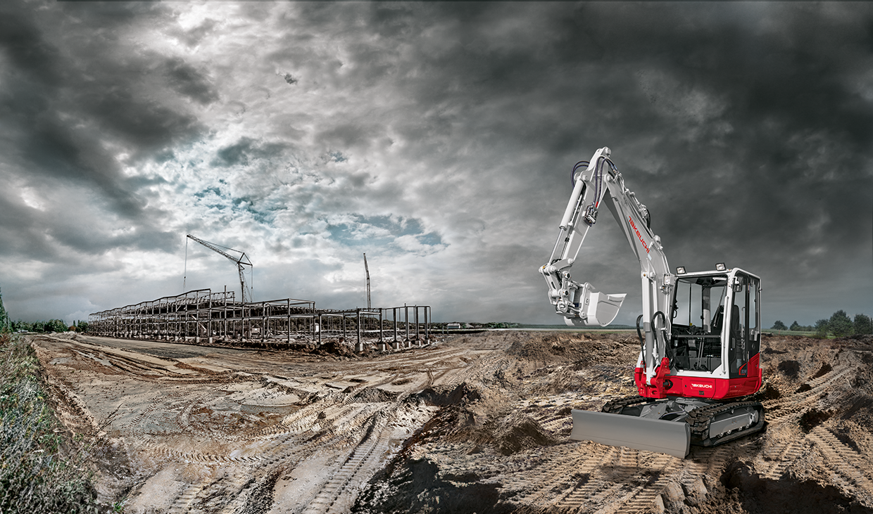 Ein rot weißer Bagger vor dunklen Wolken, im Hintergrund eine Baustelle mit Kränen