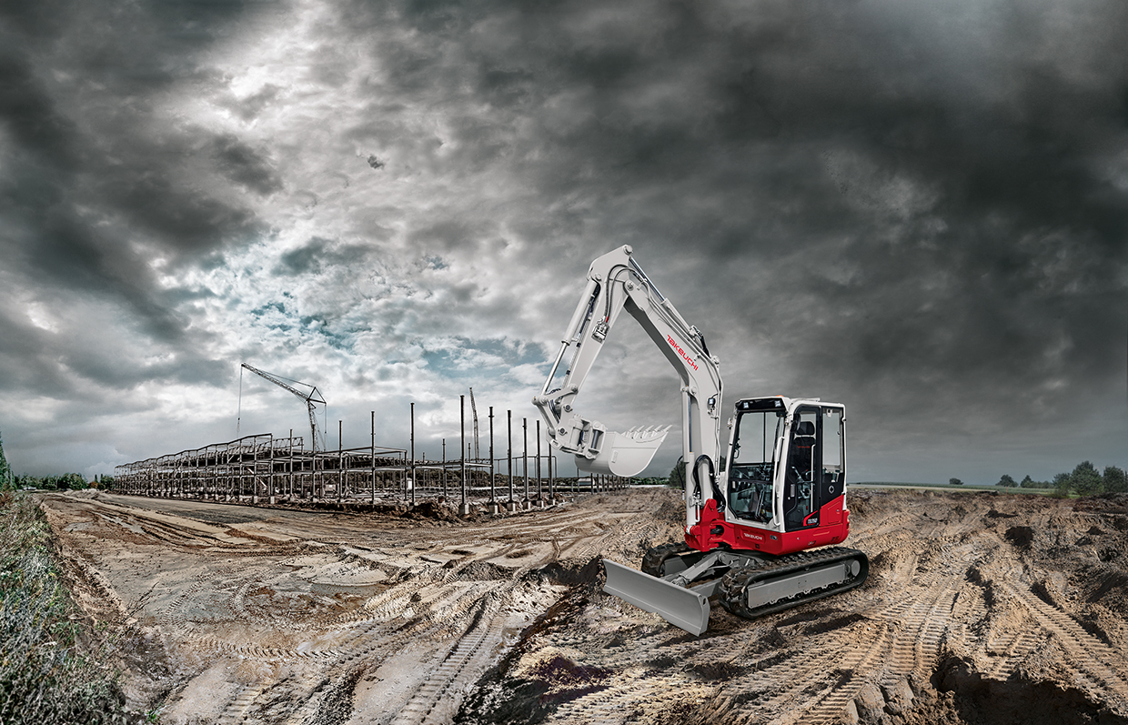 Ein rot weißer Bagger vor dunklen Wolken, im Hintergrund eine Baustelle mit Kran